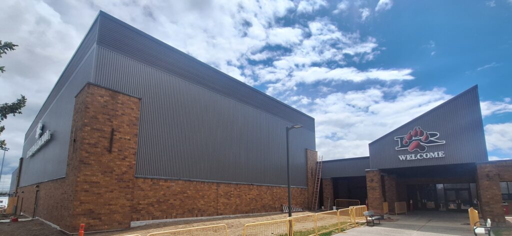 Exterior view of a large commercial building with dark gray metal siding and brick accents, featuring an angled entrance canopy with a bear paw logo and the word ‘WELCOME'.