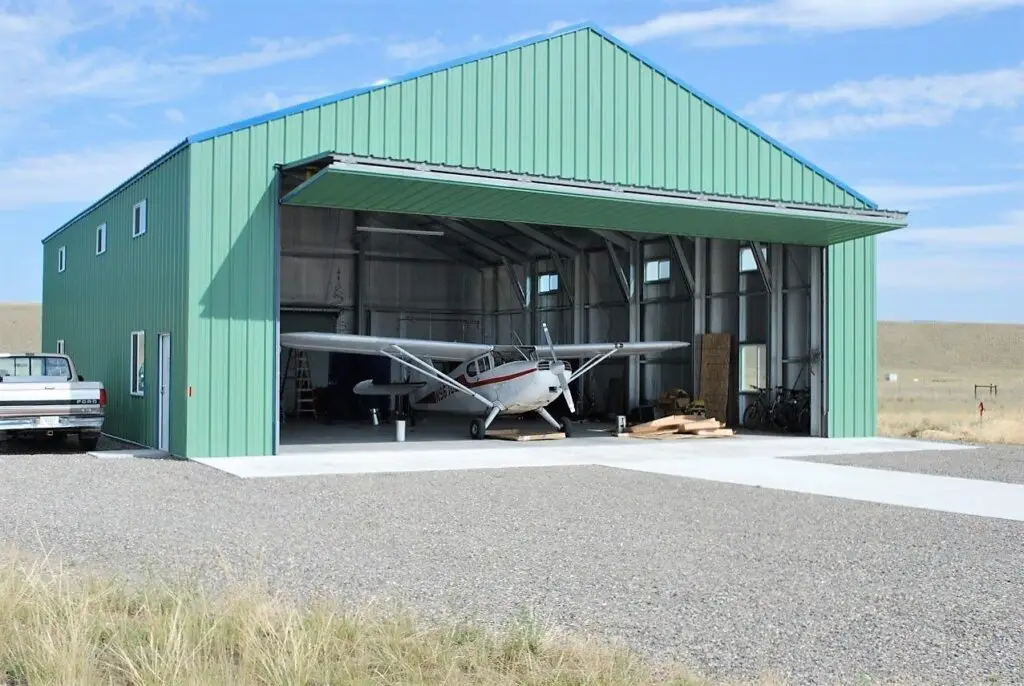 A large green metal airplane hangar with its door raised, showing a small aircraft and equipment inside.