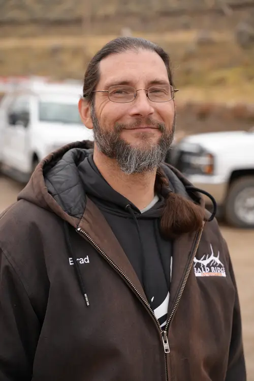 Employee wearing glasses and a brown Bald Ridge jacket smiles outdoors with white work trucks in the background.