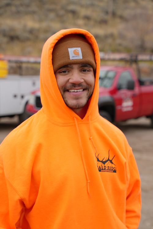 Employee wearing an orange Bald Ridge hoodie and brown beanie smiles outdoors with work trucks in the background.