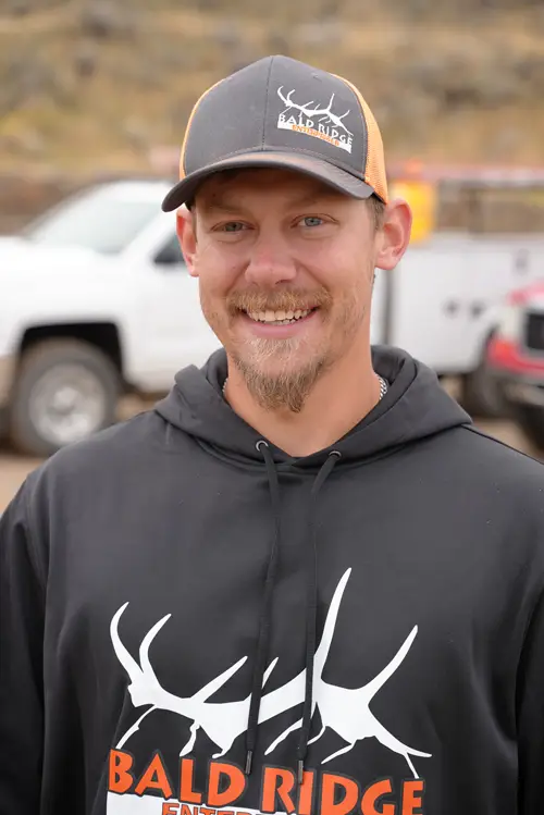 Bald Ridge Enterprises employee wearing a black company hoodie and gray cap, smiling in front of work trucks at an outdoor jobsite.