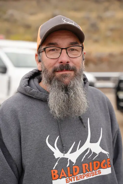 Bald Ridge Enterprises employee with glasses and a long gray beard wearing a gray company hoodie, standing outdoors near white work trucks.