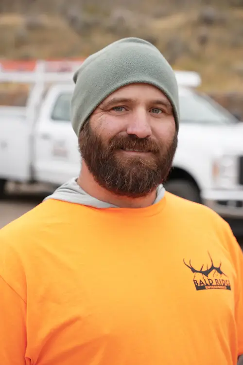 Bald Ridge Enterprises employee wearing a gray beanie and bright orange company shirt, standing outdoors in front of a white work truck.