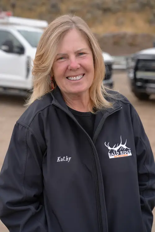 Bald Ridge employee in a black quarter-zip jacket and cap smiling outdoors near work trucks.