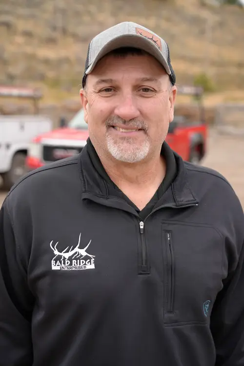 An employee wearing a black Bald Ridge Enterprises pullover and a baseball cap smiles outdoors with company trucks in the background.