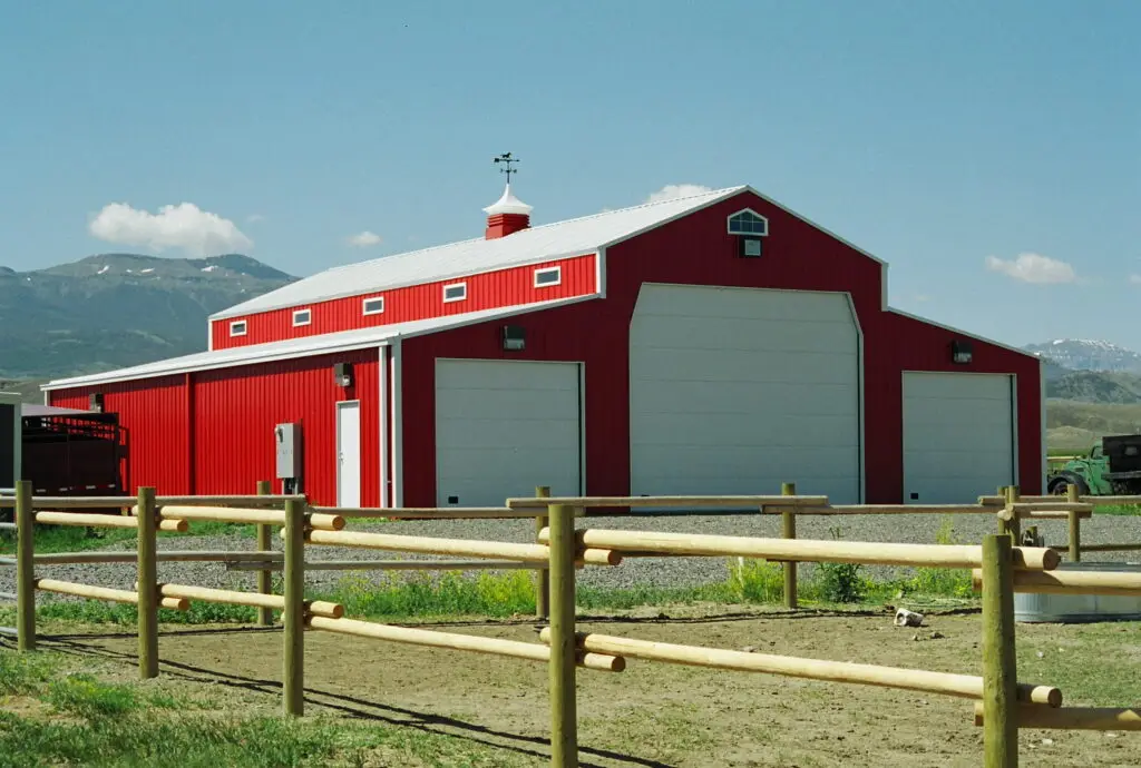 A bright red metal barn-style building with a white roof and large garage doors, set against a rural mountain backdrop.