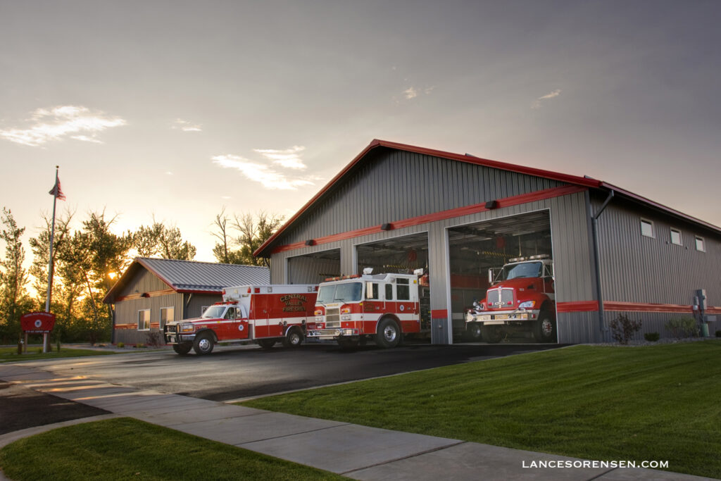 A gray and red metal fire station with multiple emergency vehicles parked outside at sunset, including fire trucks and an ambulance.