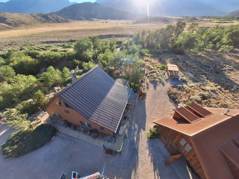Aerial view of a cabin with a dark metal roof set in an open valley with trees, fields, and mountains in the background.