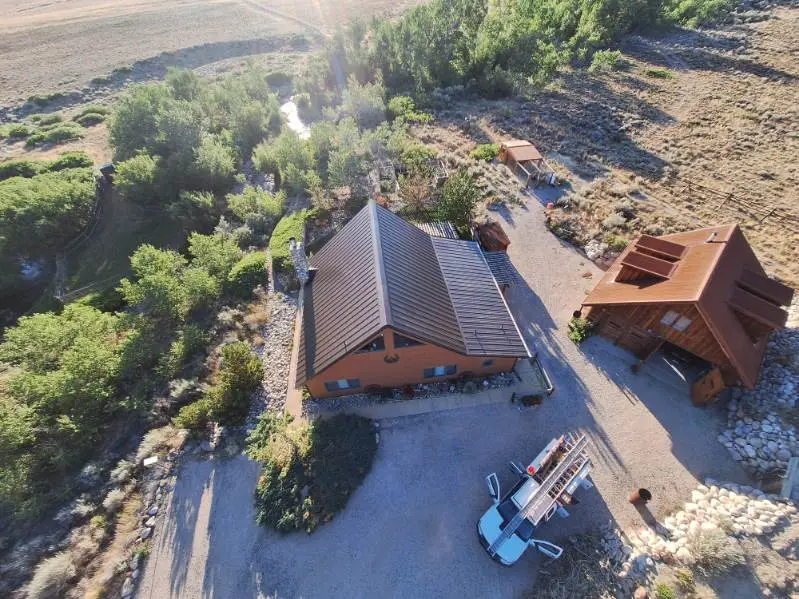 Aerial view of a cabin with a dark metal roof surrounded by trees and open landscape.