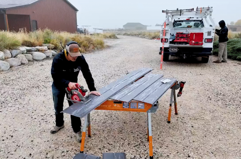 Worker cutting wood-grain siding panels on sawhorses beside a company truck on a gravel jobsite.